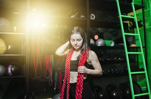 Mulher jovem muscular em sportswear com uma corda em torno de seu pescoço posando no ginásio moderno para treinamento funcional - Foto, Imagem