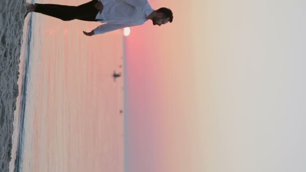 Vídeo vertical de un hombre en pantalones y una camisa caminando por la orilla del mar al amanecer. - Imágenes, Vídeo
