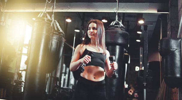 Menina atlética atraente em sportswear detém halteres em suas mãos e treina bíceps no ginásio de boxe. - Foto, Imagem