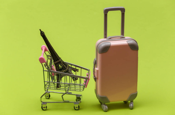 Концепция путешествий и магазинов. Мини-пластиковый дорожный чемодан и тележка супермаркета со статуэткой Эйфелевой башни на зеленом фоне - Фото, изображение