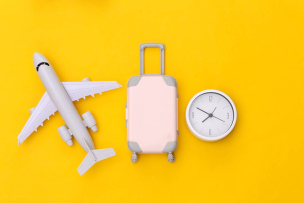 Tempo per viaggiare. Mini valigia da viaggio in plastica e orologio, aereo su sfondo giallo. Vista dall'alto. Posa piatta - Foto, immagini