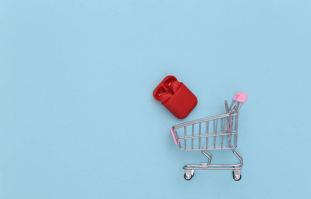 Mini shopping cart and wireless headphones in case on blue background - Foto, imagen