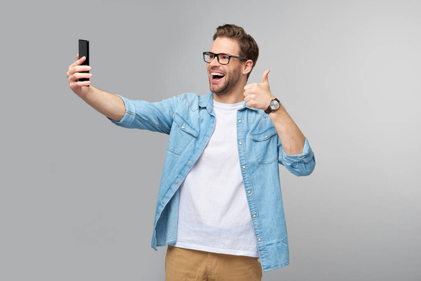 Smiling young man wearing jeans shirt taking selfie photo on smartphone or making video call standing over grey studio background - Fotografie, Obrázek