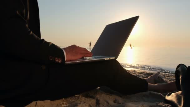 Een zakenman werkt op een laptop, zittend in een pak op het zand aan zee in de vroege ochtend. - Video