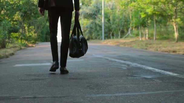 A man in a suit and bag in hand walks along the road in the park in the early morning, the camera captures the man from the back. - Footage, Video