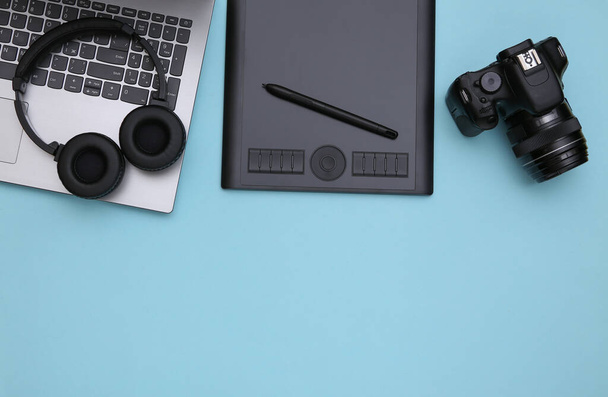 Arbeitsbereich eines Fotografen oder Retuschierers. Laptop, Grafik-Tablet, Kamera und Kopfhörer auf blauem Hintergrund. Ansicht von oben. Kopierraum - Foto, Bild