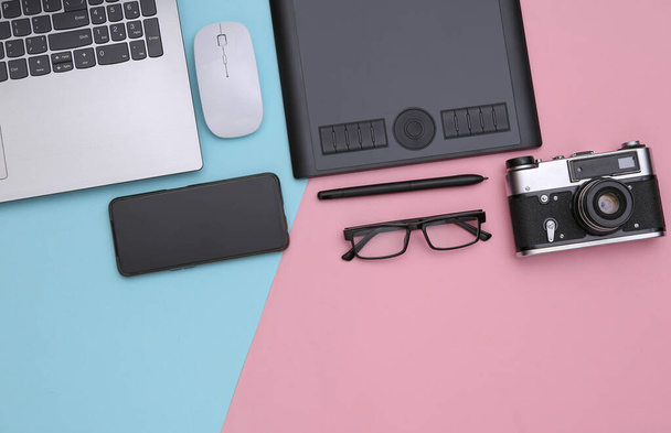 Werkruimte van fotograaf of retoucher. Laptop, grafische tablet, camera, smartphone op roze blauwe achtergrond. Bovenaanzicht. - Foto, afbeelding