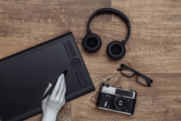 Menequin's hand works on a graphics tablet. Headphones and camera on wooden background - Photo, image