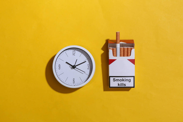 Пачка сигарет и часы на желтом фоне. Вид сверху - Фото, изображение