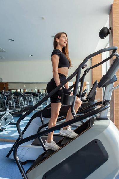 Mujer en forma sexy en ropa deportiva haciendo ejercicio cardiovascular en la máquina de ejercicios pasos en el gimnasio moderno con grandes ventanas - Foto, imagen