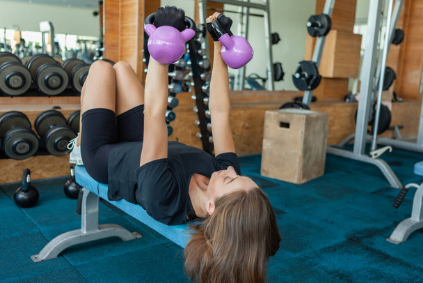 Νεαρή γυμνάστρια προπονεί θωρακικούς μυς με κουδούνια στα χέρια της ενώ ξαπλώνει σε ένα παγκάκι στο γυμναστήριο - Φωτογραφία, εικόνα