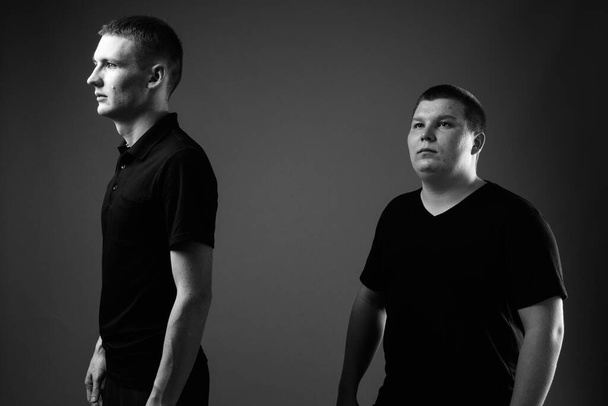 Studio shot of young man and overweight young man together against black background in black and white - Foto, afbeelding