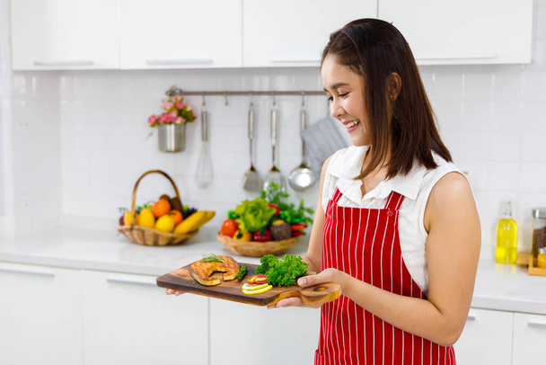 Asiatische Köchin in roter Schürze mit Lachssteak, serviert auf Holzbrett. Konzeptfrau bereitet Mahlzeiten zu Hause zu. - Foto, Bild