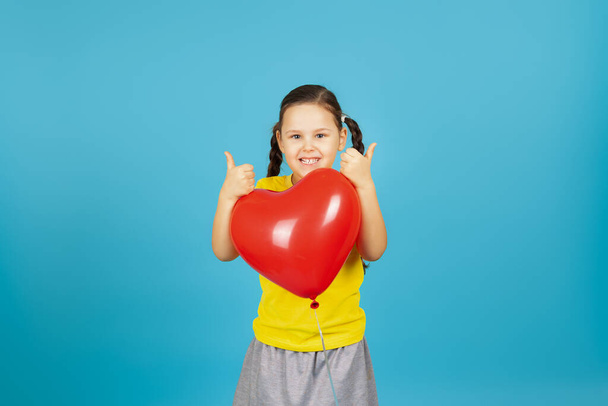 primer plano alegre, alegre pigtailed chica en una camiseta amarilla abraza un globo en forma de corazón rojo y da un pulgar hacia arriba aislado sobre un fondo azul - Foto, Imagen