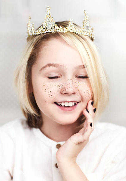 portret van mooie vrolijke androgyne jongen draagt tiara, gouden sproeten en nagellak, met lang blond haar - Foto, afbeelding