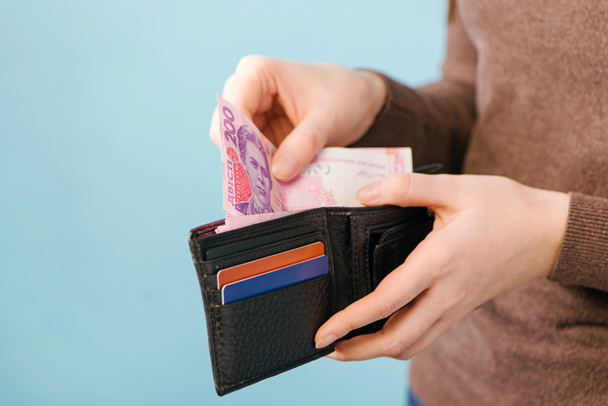 Женщина вынимает украинские гривневые деньги из сумочки на синем фоне с местом для текста. Концепция оплаты. - Фото, изображение