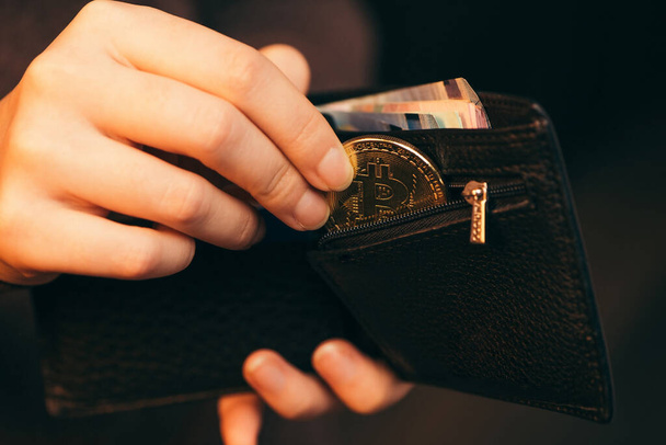 Hand putting golden bitcoin into her wallet on dark background, close up. - 写真・画像