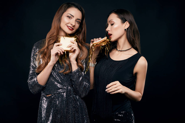 Portrait of two pretty crazy best friends in fashion dresses eating sandwich with Christmas garlands on black background in studio - Photo, Image