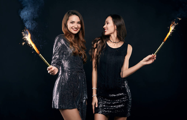 Portrait de deux assez fous meilleurs amis en robes de mode posant avec des feux d'artifice sur fond noir en studio - Photo, image