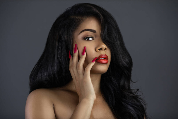 Un retrato de una joven latina con el pelo largo y ondulado negro, hermoso maquillaje, labios rojos y uñas posando sola en un estudio con un fondo gris. - Foto, imagen