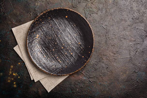 Top view of empty black ceramic plate with linen napkin on dark brown concrete backgrownd with copy space - Zdjęcie, obraz