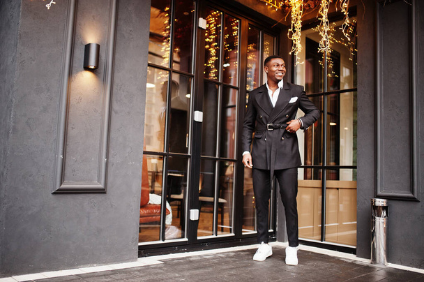 Portrait of young and handsome african american businessman in suit and white sneakers against house with garland. - Foto, Bild