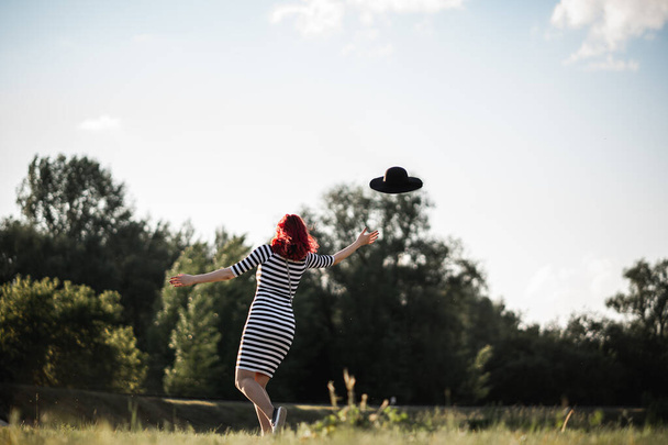 A young girl with red hair throws up her hat. Beautiful green landscape with trees and a sunlit meadow. Having fun in nature, freedom, walking or vacation concept. - Photo, Image