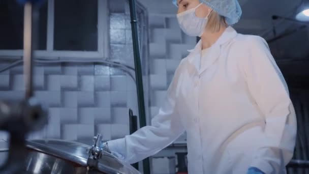 Uma mulher usando uma máscara protetora abre a tampa de um tanque de metal em uma fábrica de processamento de alimentos - Filmagem, Vídeo