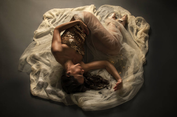 Bailarina. Jovem dançarina de balé elegante, vestida com vestido brilhante, sapatos de balé e véu transparente deitado no chão. Bela pose de balé clássico - Foto, Imagem
