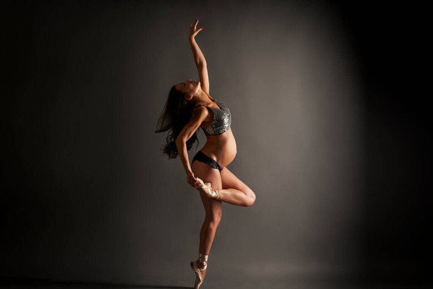 Jovem bailarina grávida realizando pose de balé clássico com top de lantejoulas e roupa interior preta em estúdio - Foto, Imagem