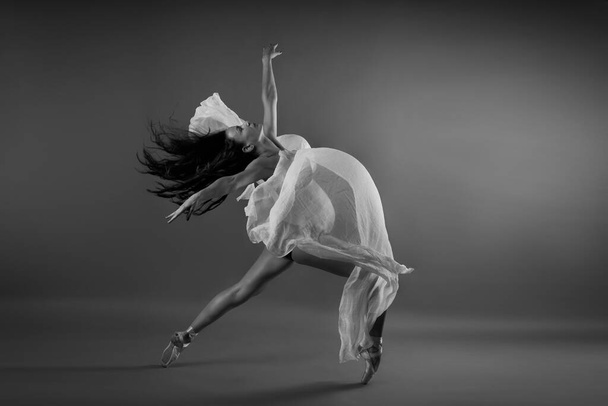 Young pregnant ballerina performing classical ballet pose with silk cloth in studio in black and white - Foto, imagen
