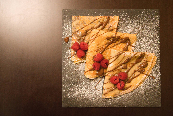 Crêpes gefüllt mit Schokolade mit Himbeeren und glasiertem Zucker auf einem quadratischen Schieferteller - Foto, Bild
