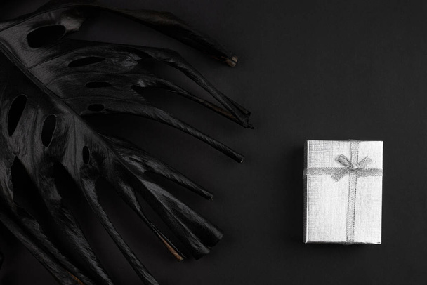 Πολυτελές μαύρο φύλλο monstera και ασημένια συσκευασία δώρου πάνω από σκούρο ματ φόντο. χώρος για κείμενο - Φωτογραφία, εικόνα