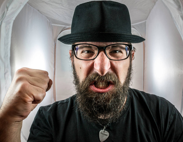 Un hombre de aspecto barbudo y enojado, parecido a un artista o músico con gafas negras y sombrero mostrando puño - Foto, Imagen