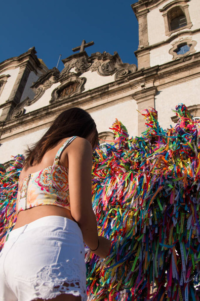Chica haciendo peticiones y atando cintas de colores en la rejilla frente a la iglesia Bonfim en Salvador Bahia Brasil. - Foto, Imagen