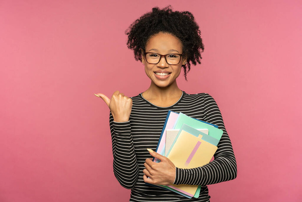 Afroamerikanische Tutorin oder Lehrerin isoliert an rosa Studiowand. Studentin trägt Brille, Notizbücher in der Hand, lächelt und zeigt Finger auf freien Raum. Ausbildung in der High School University College. - Foto, Bild