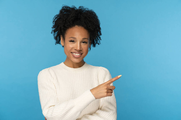Smiling dark skinned woman with curly hair wear white jumper pointing with finger, showing blank copy space for advertising, offering, product, promotion, sale, isolated on studio blue background. - Photo, Image
