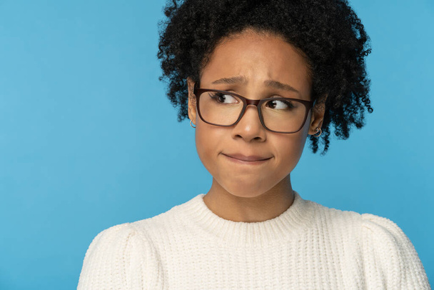 close-up studio portret van verlegen lastige jonge Afro Amerikaanse vrouw dragen bril witte trui, bijtende lippen gevoel verlegen, verward en nerveus, kijken opzij, geïsoleerd op blauwe achtergrond. - Foto, afbeelding