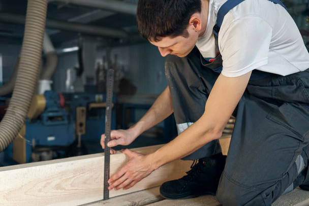 Młody człowiek pracuje w stolarni. Pracownik mierzy drewno linijką - Zdjęcie, obraz