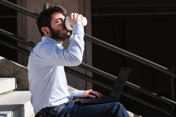 Молодой бизнесмен пьет кофе, сидя на лестнице с ноутбуком. Бизнесмен на городской улице города. - Фото, изображение