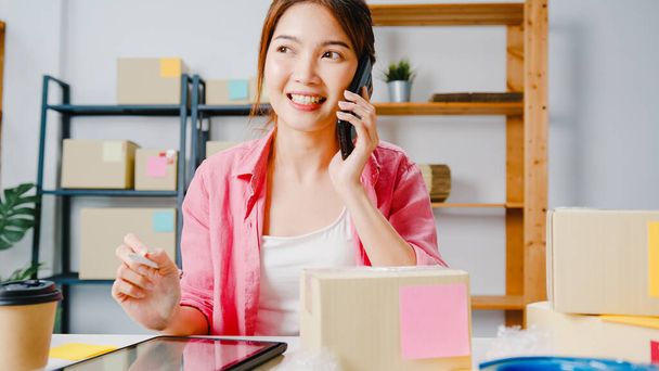 Young Asia businesswoman using mobile phone call receiving purchase order and check product on stock, work at home office. Small business owner, online market delivery, lifestyle freelance concept. - Photo, Image