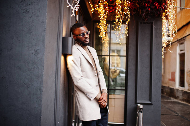 Stylish african american man wear beige jacket with sunglasses pose against house with golden garland. - Foto, immagini