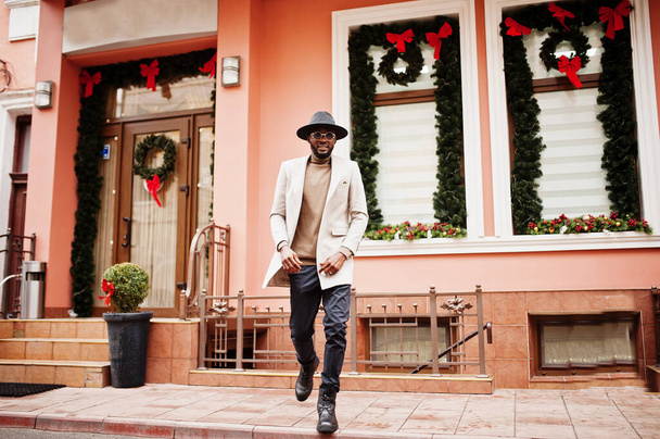 Stylish african american man wear beige jacket and  black hat with sunglasses pose against house with new year decorations and wreath. - Фото, зображення