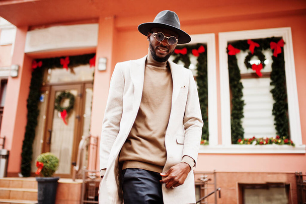Stylish african american man wear beige jacket and  black hat with sunglasses pose against house with new year decorations and wreath. - Foto, Imagen