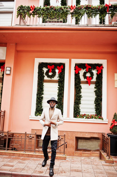 Stylish african american man wear beige jacket and  black hat with sunglasses pose against house with new year decorations and wreath. - Foto, Bild