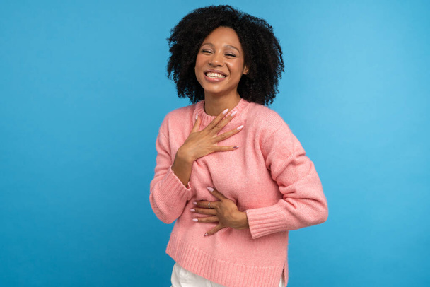 Lachende gemengde ras millennial vrouw met krullend haar in casual roze trui, houdt de hand op de borst, plezier hebben, geïsoleerd op studio blauwe achtergrond. Een dolblij Afrikaans meisje dat glimlacht. Positieve emoties. - Foto, afbeelding