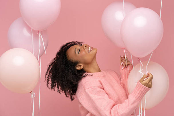 Happy pleased biracial woman with curly hair closed eyes, holding a lot of balloons, enjoys cool party, wears pink sweater, celebrates birthday, standing over studio pink background. Festive event - Foto, imagen