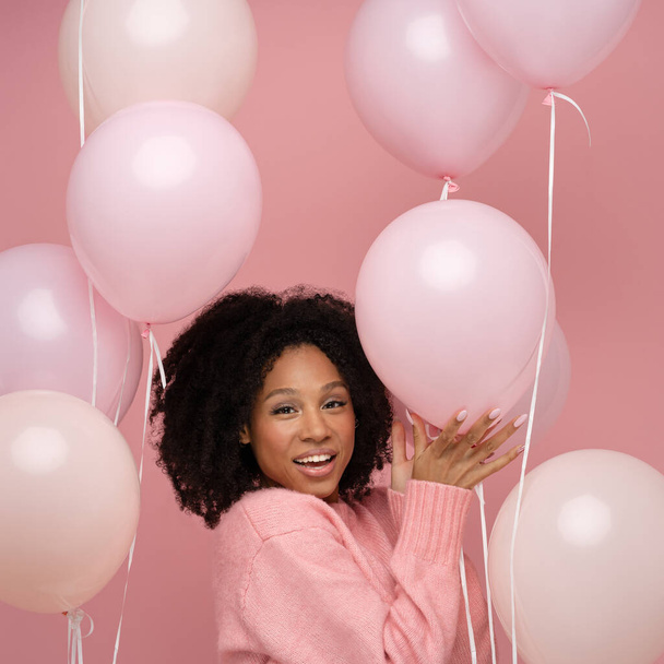 Happy pleased biracial woman with curly hair holding a lot of balloons, enjoys cool party, wears pink sweater, celebrates birthday, look at camera, standing over studio pink background. Festive event - Photo, image