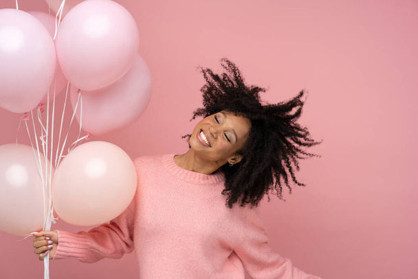 Happy pleased biracial woman with curly hair dancing, holding many balloons, enjoys cool party, wears pink sweater, celebrates birthday, standing over studio pink background. Copy space, advertising. - Foto, Bild