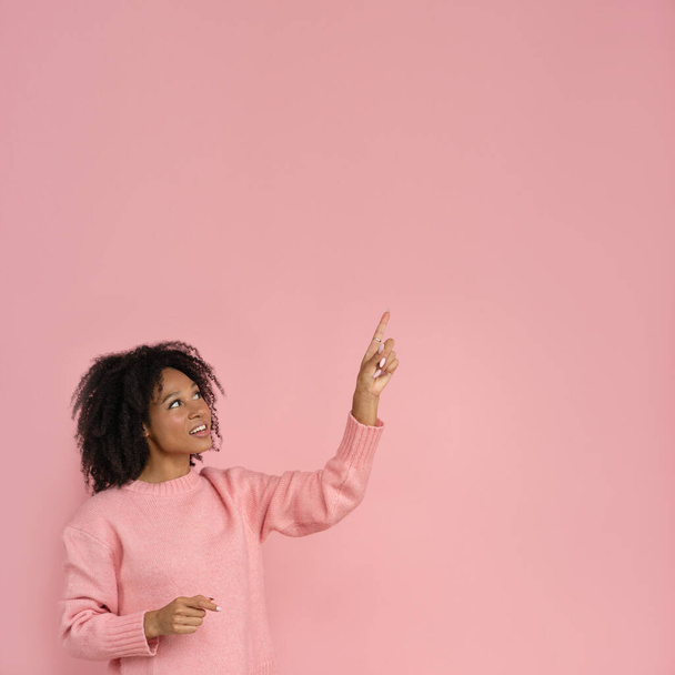 Smiling dark skinned woman with curly hair wear pink jumper point up with finger, showing blank copy space for advertising, offering, product, promotion, sale, isolated on studio pink background. - Photo, Image